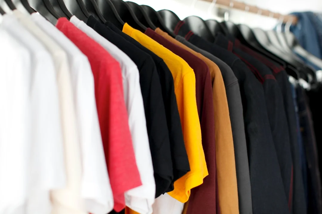 A neatly folded stack of white and neutral t-shirts in varying weights arranged on a light wood surface, with a few unfolded tees draped naturally in the background